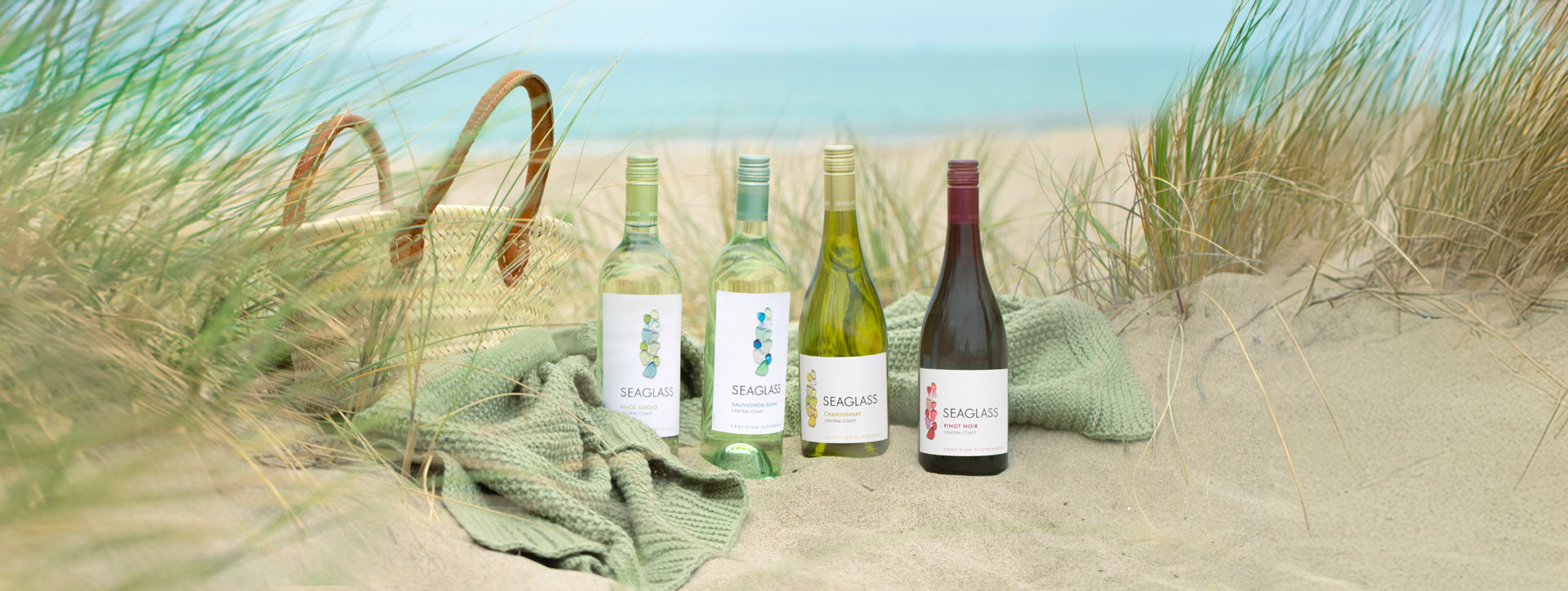 Four bottles of Seaglass wine on a sand dune next to a wicker bag, surrounded by beach grass, with a blue sea in the backgroun