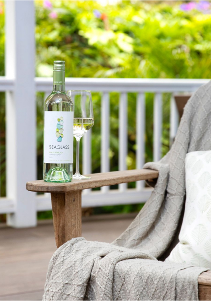 A bottle of Seaglass pinot grigio white wine on the arm of a wooden chair on a porch with white railings and green plants in the background