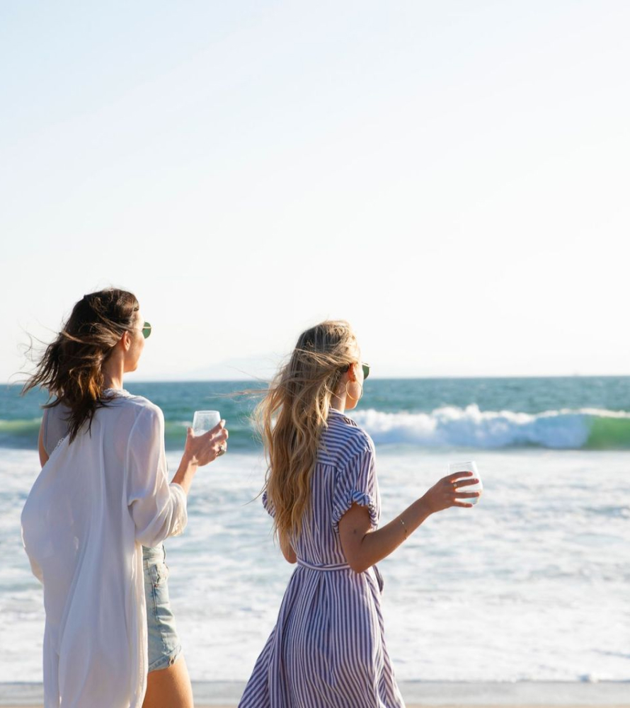 Two women wearing light-colored summer dresses drink wine on the beach with waves and blue sky in the background