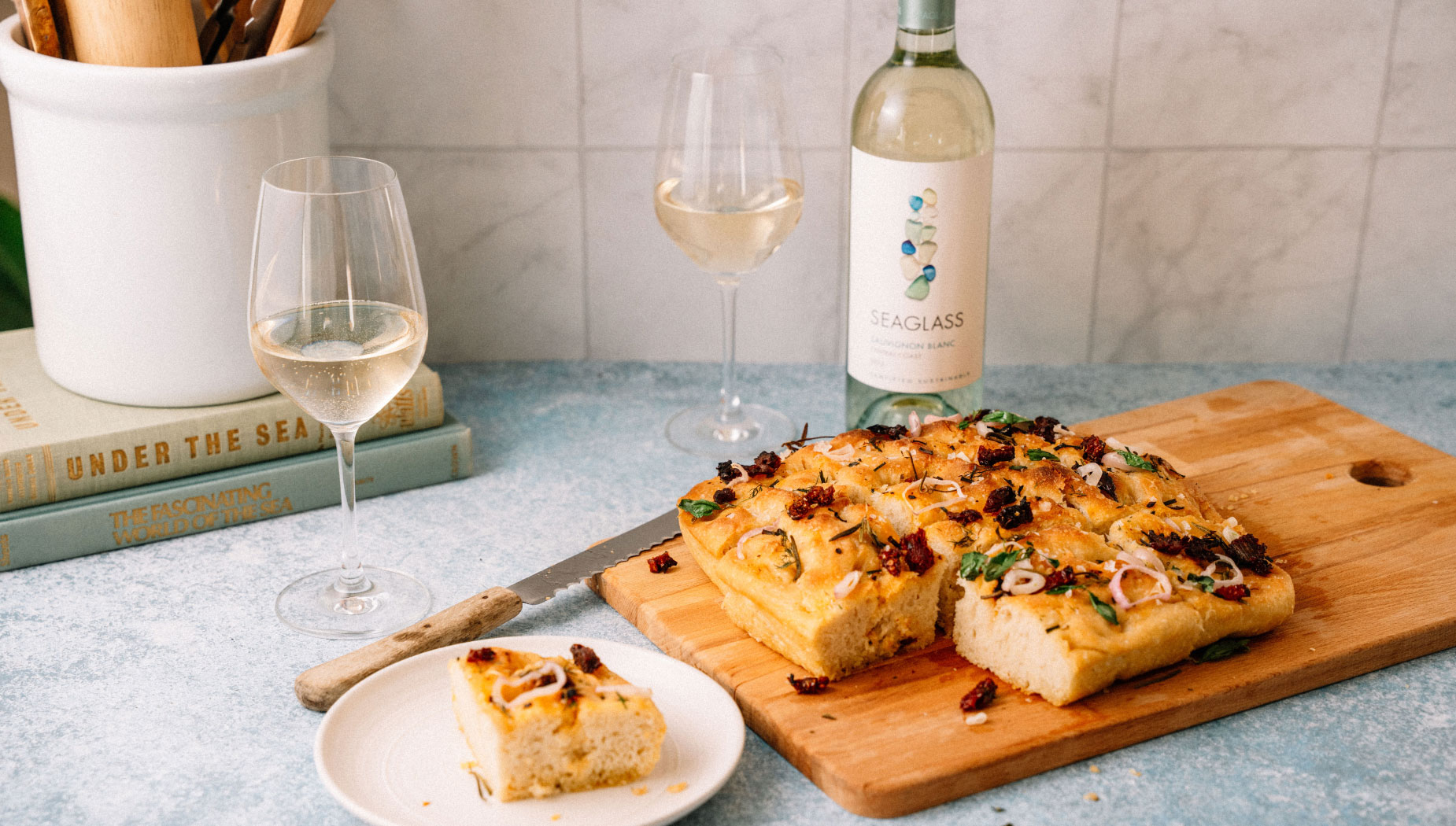 Sliced focaccia lies on a wooden cutting board, next to a bread knife, two glasses of white wine, and a bottle of Seaglass Central Coast Sauvignon Blanc