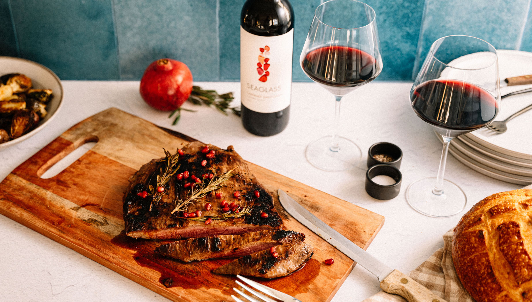 Sliced flank steak lies on a wooden chopping board next to a whole pomegranate, a sprig of rosemary, a wine glass, and a bottle of SEAGLASS Cabernet Sauvignon