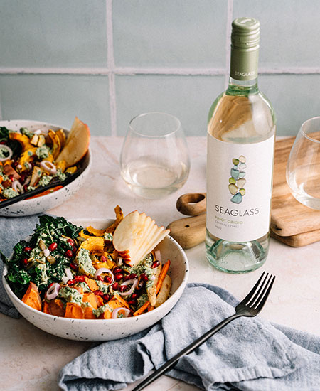 A harvest bowl on a table with a blue linen napkin, two stemless glasses of white wine, a wooden chopping board, and a bottle of SEAGLASS Central Coast Pinot Grigio
