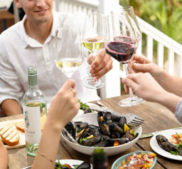 A man in a white shirt holds up his wine glass to toast the glasses of several other people who are mostly out of frame over a dinner table with plates of seafood and bread, and a large bowl of mussels, as well as a bottle of Seaglass sauvignon blanc